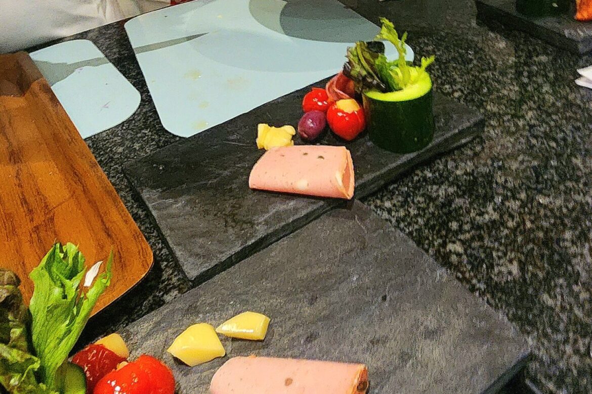 At the Singles Cooking Club event, people in aprons prepare a charcuterie board with assorted meats, cheeses, grapes, and leafy greens on black serving plates.
