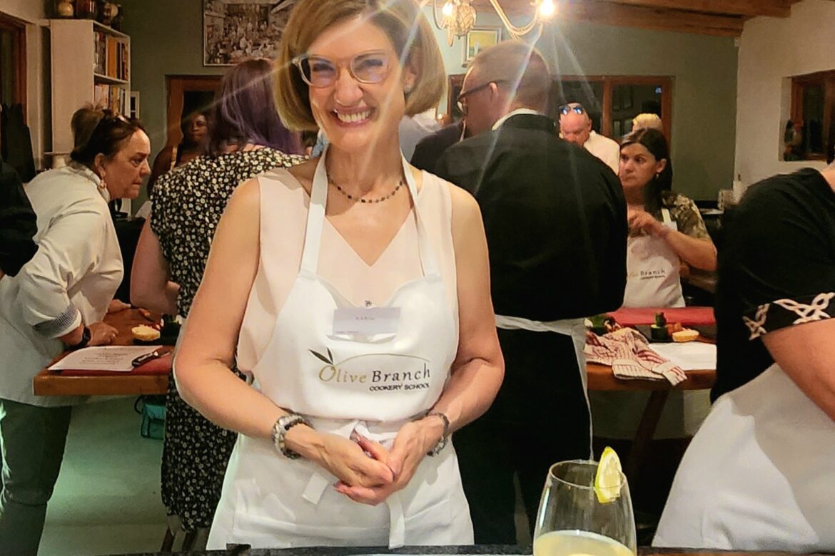 A woman in glasses and an apron beams at the Singles Cooking Club, surrounded by fellow participants in a charming kitchen. Ingredients and a refreshing drink with lemon adorn the table before her, while wooden beams and a chandelier add elegance to this lively singles event.
