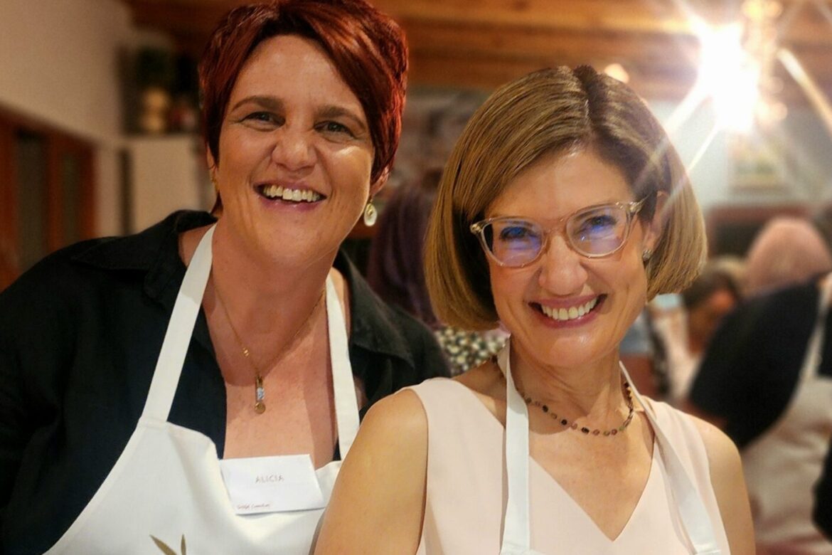 Two women, both smiling in white aprons at a Singles Cooking Club event, pose for the camera. One has short red hair, and the other sports short blonde hair and glasses. They stand in a warmly lit room with wooden ceiling beams behind them.