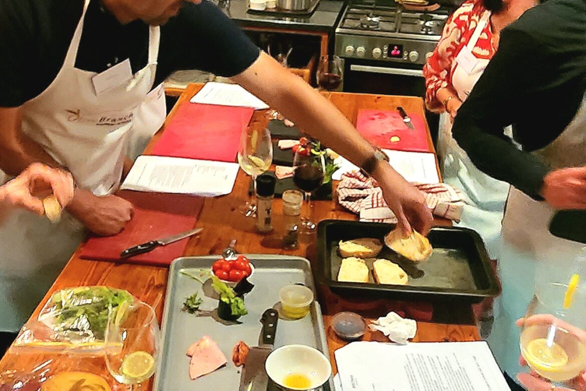 In the bustling kitchen, members of the Singles Cooking Club gather around wooden tables, preparing food. Ingredients like bread, cheese, and vegetables are spread out as two aproned individuals focus on their tasks while others chat and sip from glasses.