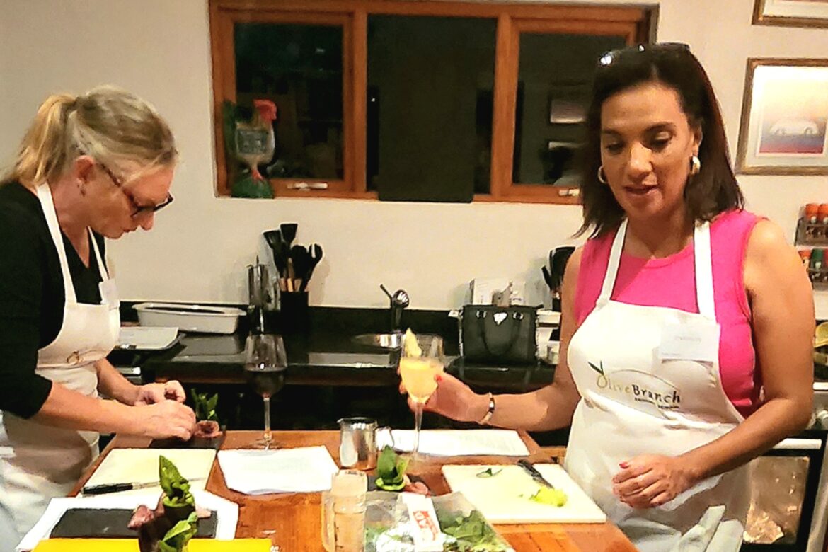 Two women in aprons are preparing food in a cozy kitchen, part of their Singles Cooking Club gathering. One chops ingredients while the other holds a drink. The countertop is filled with various cooking items, and the background reveals a window and neatly arranged kitchen utensils.