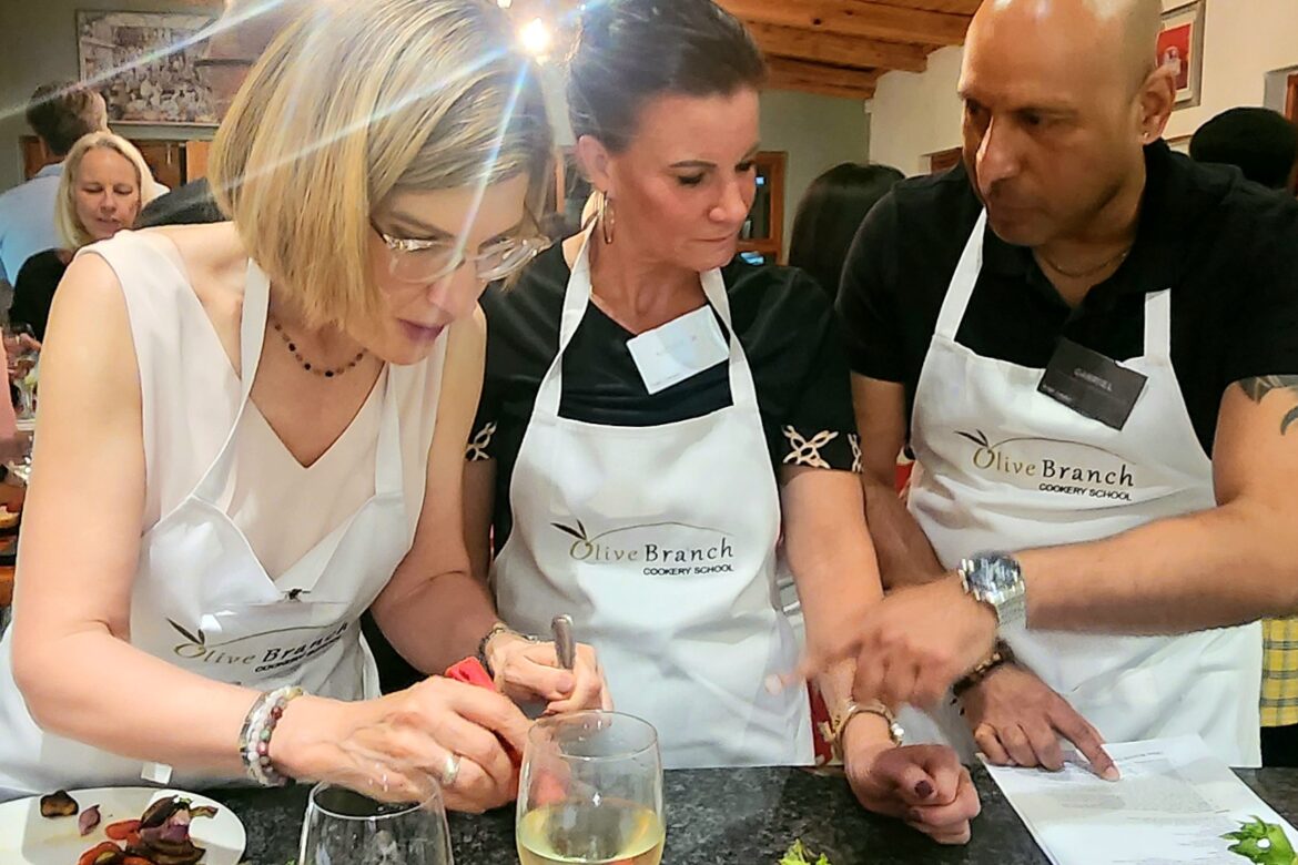 Three people wearing aprons gather around a kitchen counter at a Singles Cooking Club, preparing food together. Two wine glasses and a plate with vegetables accompany the toasted bread. The lively setting suggests an engaging cooking workshop perfect for meeting others who share your culinary passion.