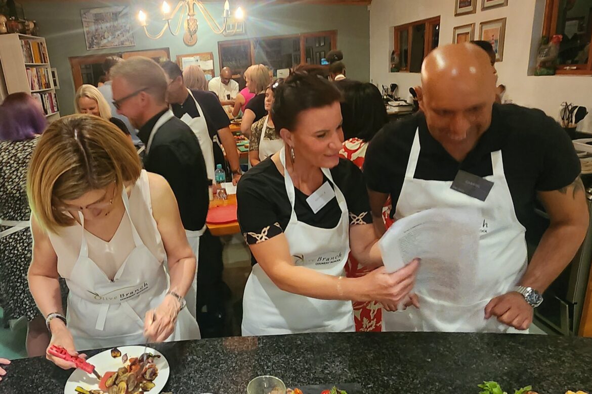 A group of singles wearing aprons participates in a lively cooking class. Two are focused on plating dishes, while the others chat animatedly in the background. A variety of ingredients and drinks are spread across a black counter, enhancing this engaging Singles Cooking Club experience.