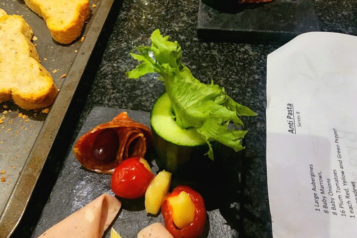 A Singles Cooking Club gathering showcases a kitchen counter laden with sliced bread, cured meats, cheese, cherry tomatoes, cucumber, and lettuce on sleek black slate boards. In the background, a wine glass and additional prepared dishes hint at the evening's delectable single events.