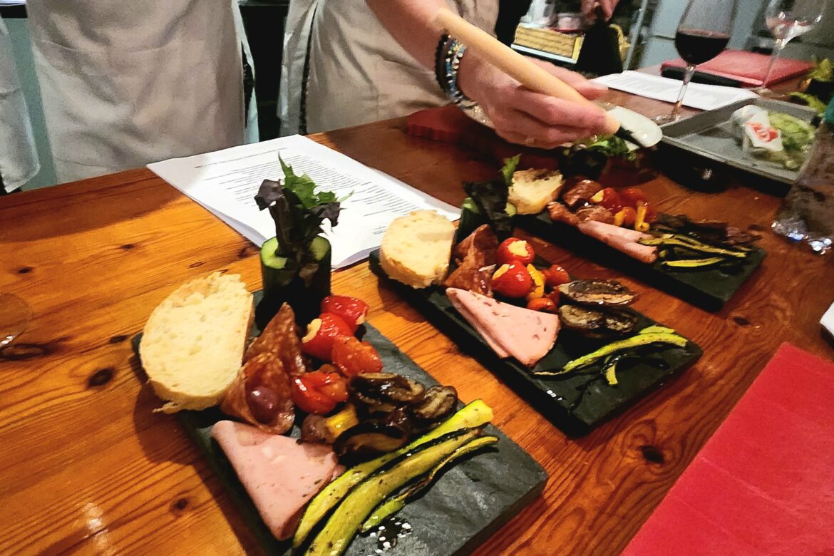 Three slate plates laden with assorted meats, cheese, grilled vegetables, and bread slices are served on a wooden table at the Singles Foodies Event. Hands of a person in an apron attend to the dishes while glasses of red wine set the mood in the background.