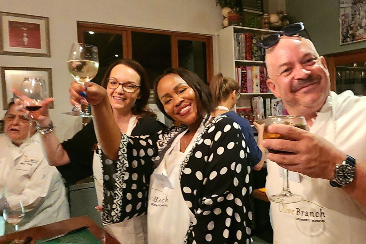 In a warm and inviting kitchen, members of the Singles Cooking Club gather around the counter. Two women and two men in aprons hold wine glasses, smiling for the camera with delicious food and drinks spread before them.