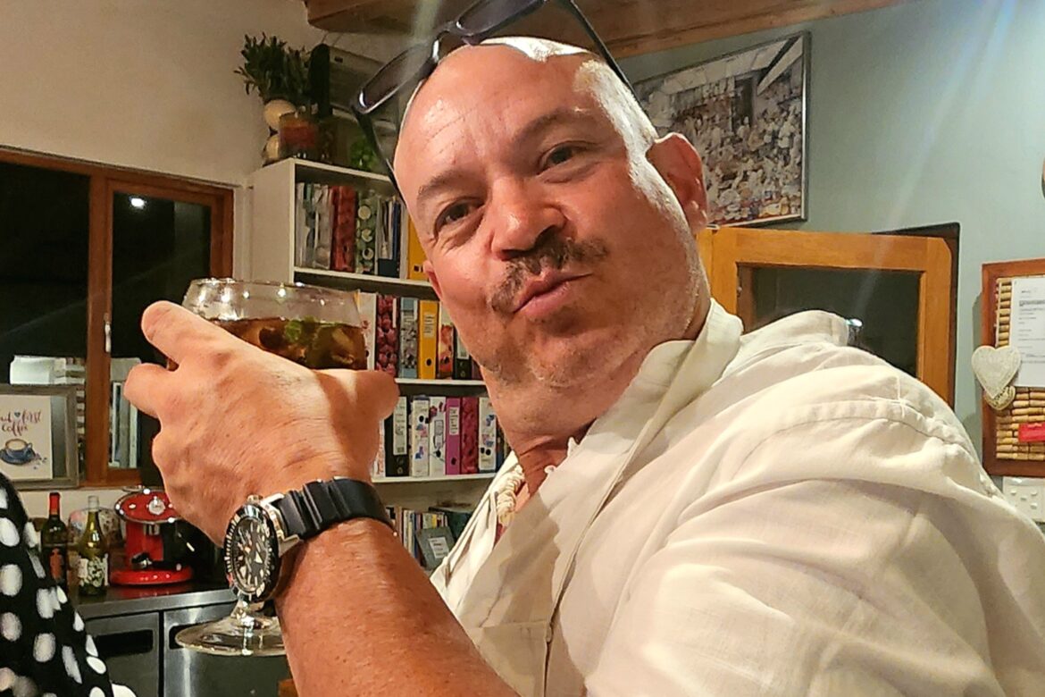 A person wearing sunglasses on their head holds a drink and poses playfully in a kitchen setting, perhaps getting ready for the Singles Cooking Club. They sport a white shirt and a watch, with kitchen shelves and a door visible in the background.