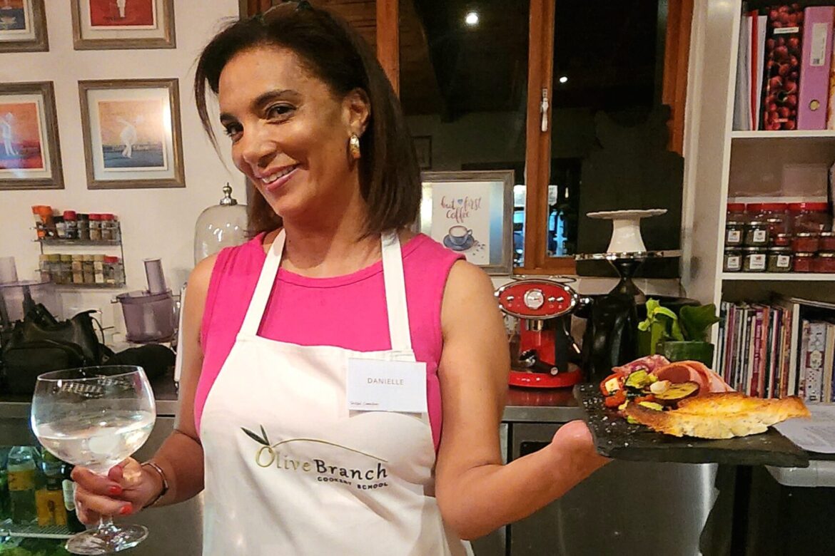 A woman wearing a white apron holds a glass of water and a platter with assorted food, including cured meats and cheese. She's smiling in a kitchen setting filled with art and shelves, perfect for the Singles Foodies Event she's hosting, where culinary enthusiasts mingle and savor delights.