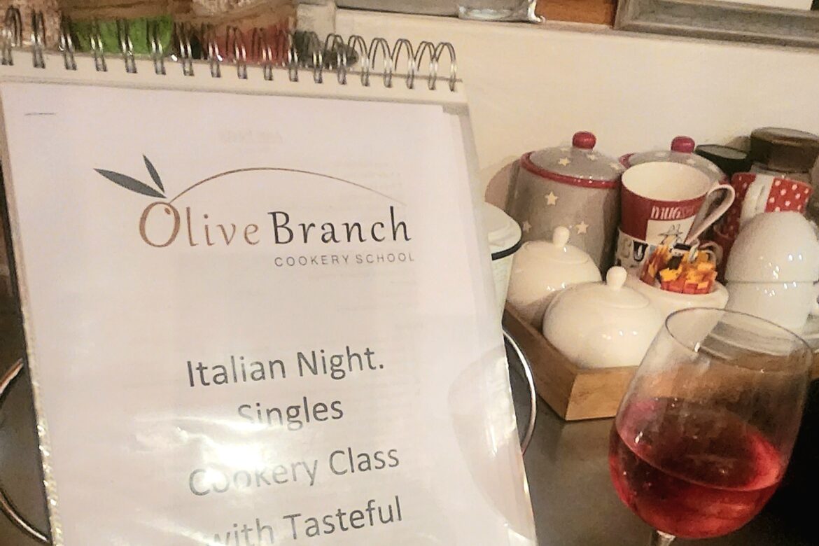 A sign on a stand reads Olive Branch Cookery School. Italian Night. Singles Cooking Club with Tasteful Connections. A glass of red wine and various kitchen items are on the counter beside it.