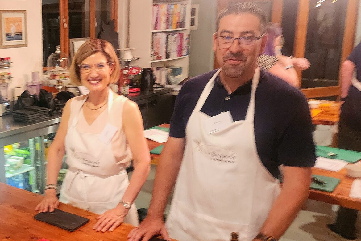 A man and a woman are in a kitchen setting, both wearing aprons. They stand side by side at a wooden counter with various kitchen tools in the background, smiling as if part of a Singles Cooking Club event.