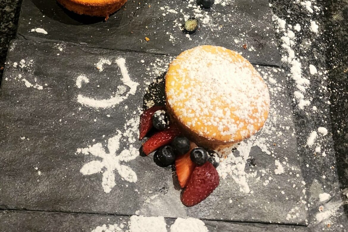 At a Singles Foodies Event, five round cakes topped with powdered sugar, sliced strawberries, and blueberries are arranged in a line on a black slate board. The board showcases intricate powdered sugar patterns, perfect for food lovers seeking sweet delight.
