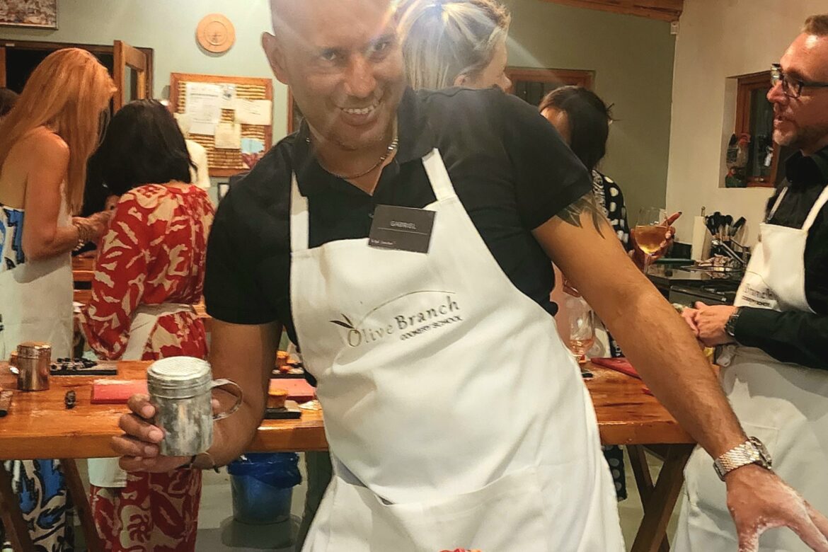 At a lively Singles Foodies Event, a participant beams in a cooking class, surrounded by beautifully decorated desserts. Clad in a white apron and wielding a shaker, they enjoy the single event while others in the background are equally engrossed in culinary adventures.