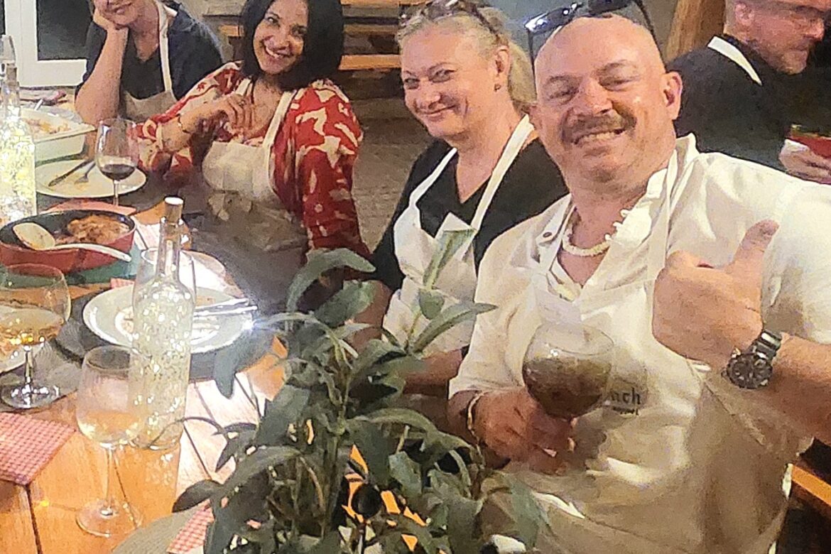 A group of four foodies at a Singles Cooking Club event sits around a wooden dining table, smiling and wearing aprons. One person gives a thumbs-up while holding a wine glass. The table is set with plates, glasses, and delicious dishes. A plant is in the foreground, adding charm to the scene.