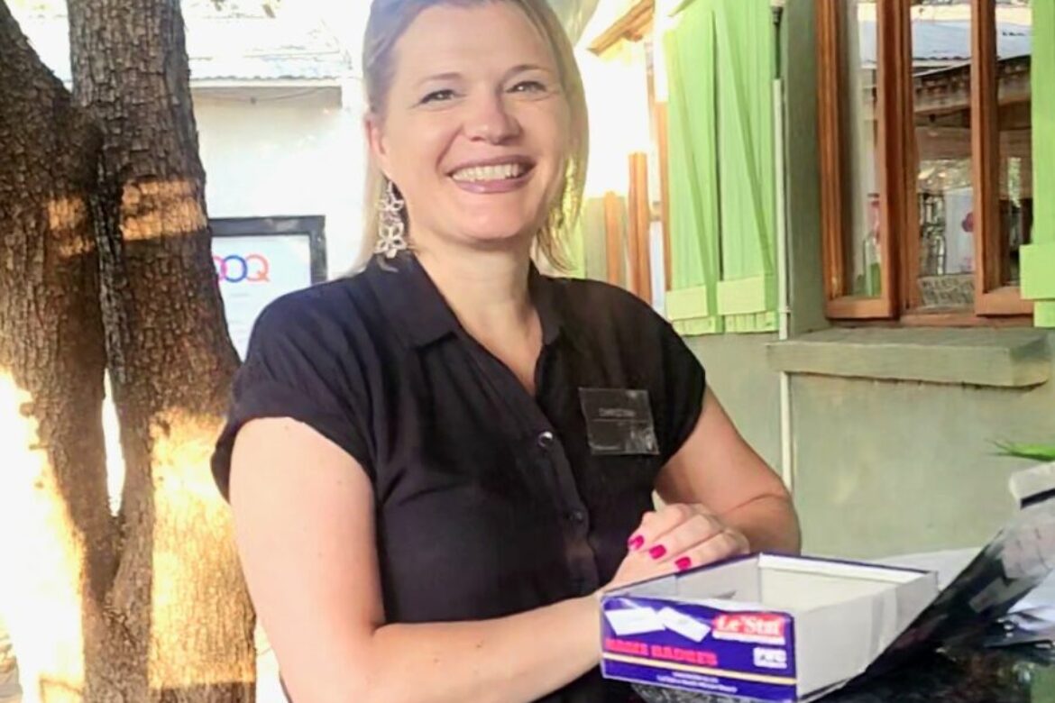 A woman in a black shirt stands outdoors next to a table with a box of envelopes, smiling at the camera. She's attending a Singles Foodies Event. In the background, there's a tree and building with green shutters, creating the perfect setting for this delightful singles gathering.