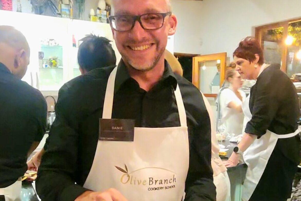 A person wearing glasses and a white apron that reads Olive Branch Cookery School smiles while holding a plate of sliced vegetables. Behind them, people bustle about in the kitchen, part of the lively Singles Foodies Event.