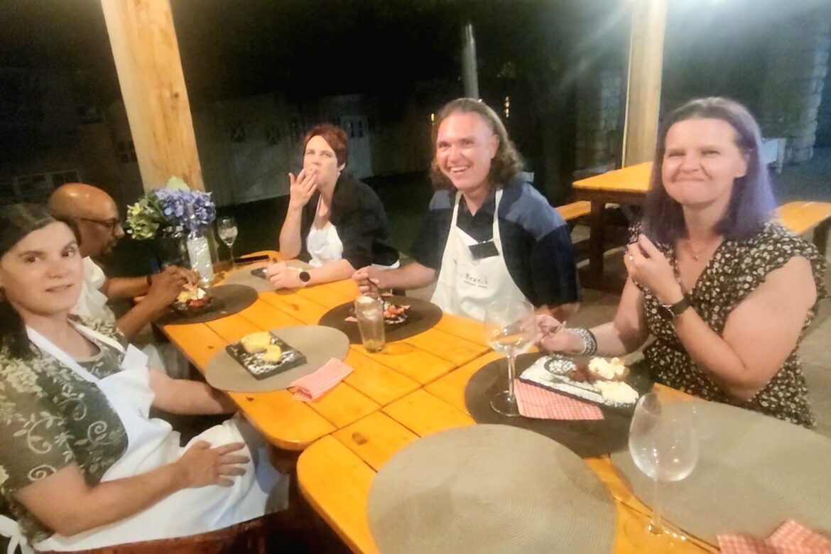 A group of five people from the Singles Cooking Club sits around a wooden table outdoors at night, enjoying a meal and smiling. The table is set with plates, napkins, and wine glasses. A vase of flowers decorates the table. They appear to be in a relaxed and happy setting.