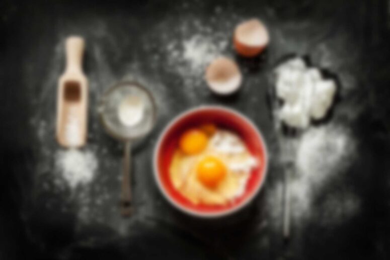 A baking setup on a dark surface for the Singles Cooking Club, featuring a bowl with eggs and flour, a cracked eggshell, a sifter, and a wooden scoop with flour. A whisk lies among scattered flour, inviting singles to mix fun and flavor together.