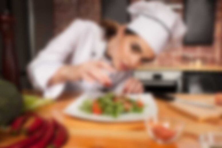 A chef in a white uniform and hat attentively garnishes a dish with greens on a plate. The background is a kitchen setting with blurred elements enhancing the focus on the chefs precise actions and the colorful dish.