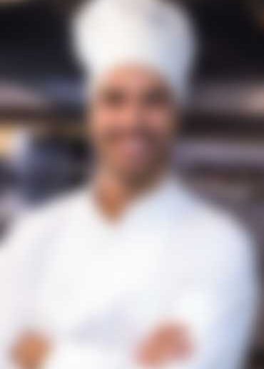 A chef wearing a white uniform and hat stands with arms crossed in a blurred kitchen background.