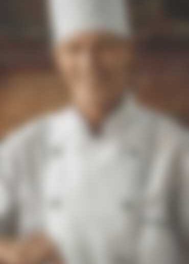 A chef in a white uniform and hat, standing in front of a brick wall. The image is blurred, obscuring details of the chefs face and expressions.