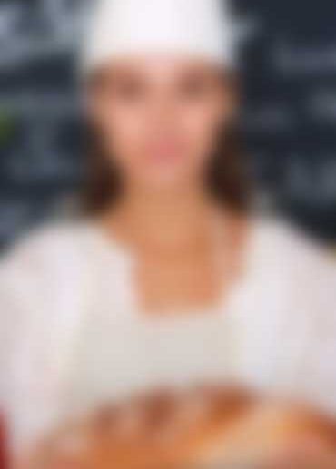 A person in a chefs hat and apron holds a freshly baked loaf of bread, standing in front of a chalkboard with blurred writing.