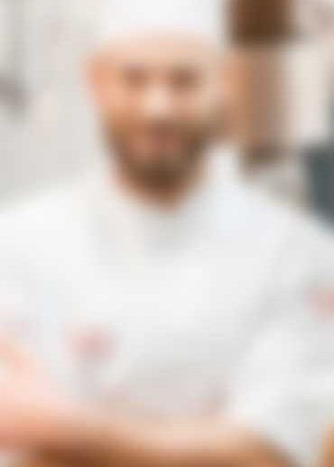 A blurred image of a person wearing a white chefs uniform and hat, standing with arms crossed. The background is indistinct, possibly a kitchen setting.