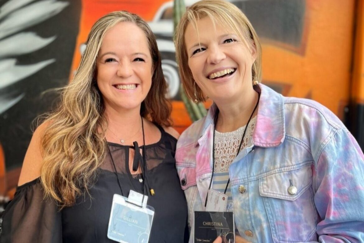 Two women smiling together in front of a vibrant, orange mural with cityscape elements are ready for the Singles Food Tour. One is wearing a black outfit and the other sports a pastel tie-dye jacket, both displaying name tags on their lanyards.