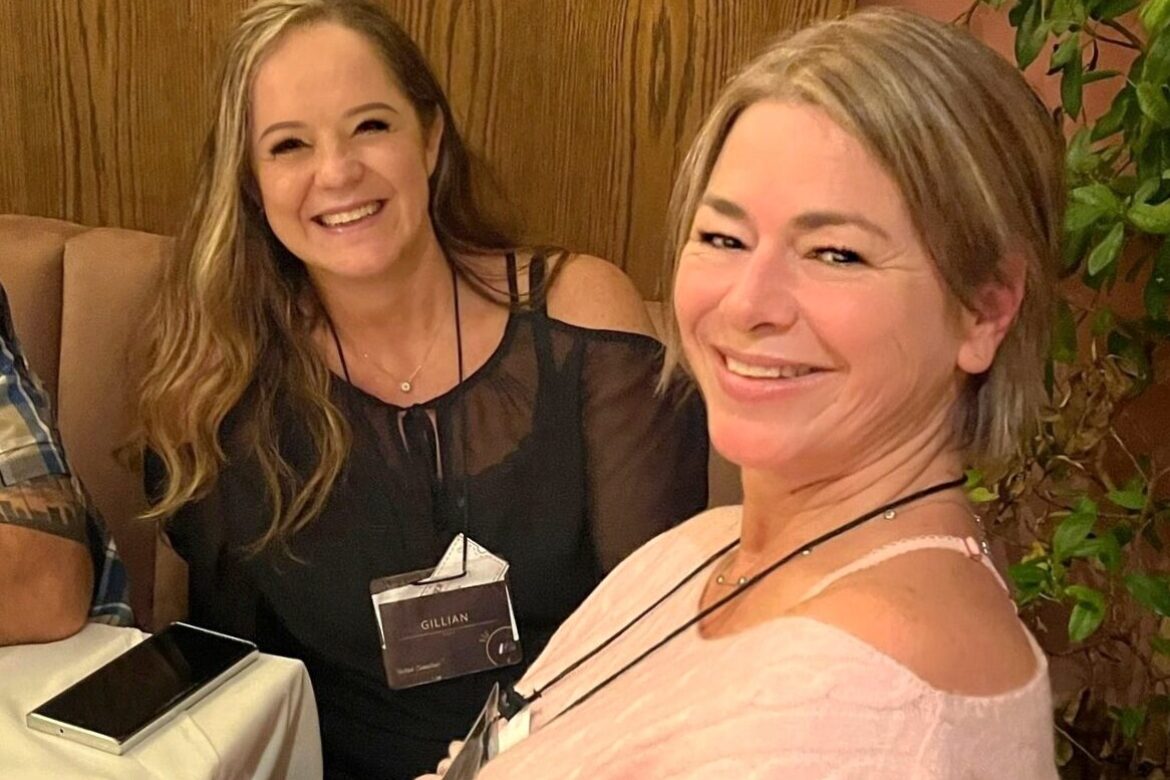 Two women are sitting at a table, smiling at the camera during their singles food tour. One is wearing a black top and the other a light pink top. A smartphone rests on the table, hinting at adventures to come, as a wooden panel and a plant add charm to the background.