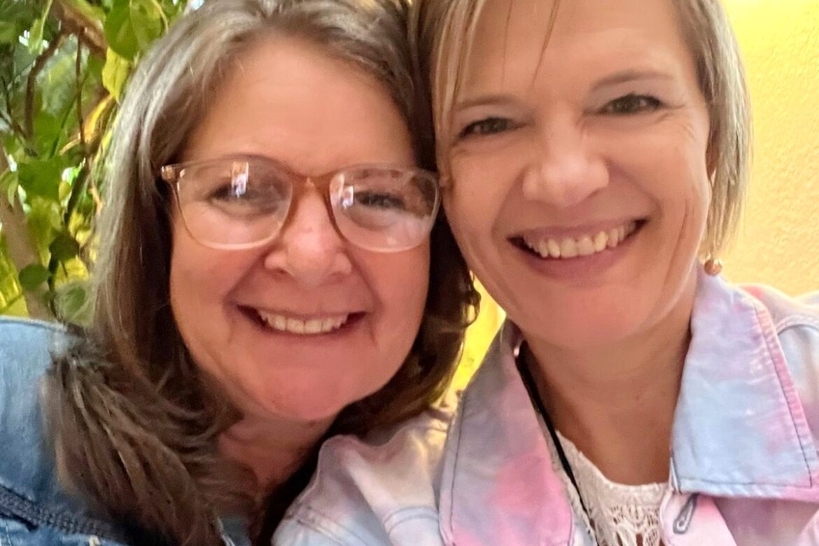 Two women beaming for a selfie indoors, capturing their joy on a Singles Food Tour. The one on the left flaunts long brown hair and glasses with a denim jacket, while her friend sports short blonde hair in a pastel denim jacket. A plant and light fixture cozy up the background.