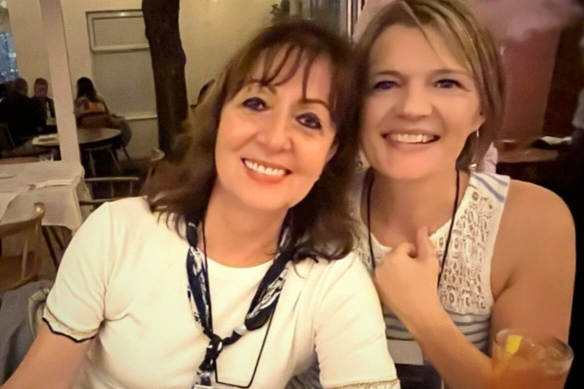 Two women seated at a restaurant table smile for the camera during their singles food tour. One wears a white top with a patterned scarf, and the other a lace white top. A drink with a lemon slice is on the table, and string lights twinkle in the background.
