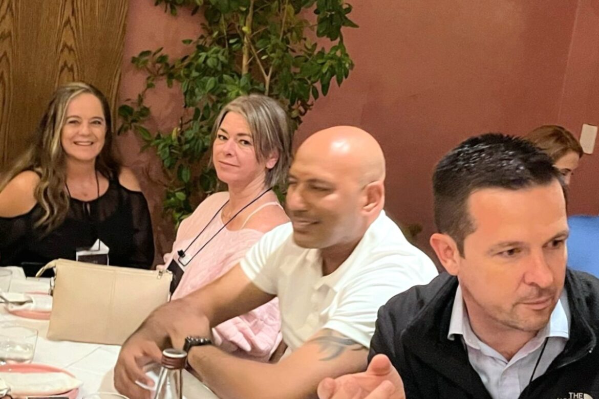 A group of four people, perhaps enjoying a Singles Food Tour, sit at a restaurant table. They are smiling and engaged in lively conversation. Plates, glasses, and a water bottle are scattered about. In the background, a plant adds life against the vibrant red wall.