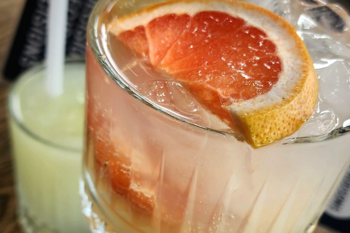 Two drinks on a wooden table capture the essence of a singles food tour. The main drink, a clear glass with ice and a slice of grapefruit, is refreshing, while the light green concoction in the background invites further exploration of flavors with each sip through its straw.