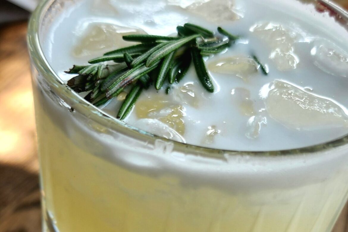 A frothy yellow cocktail in a textured glass, garnished with rosemary sprigs and ice cubes, sits enticingly on a wooden table—the perfect companion for any singles food tour under the warm glow of natural light.