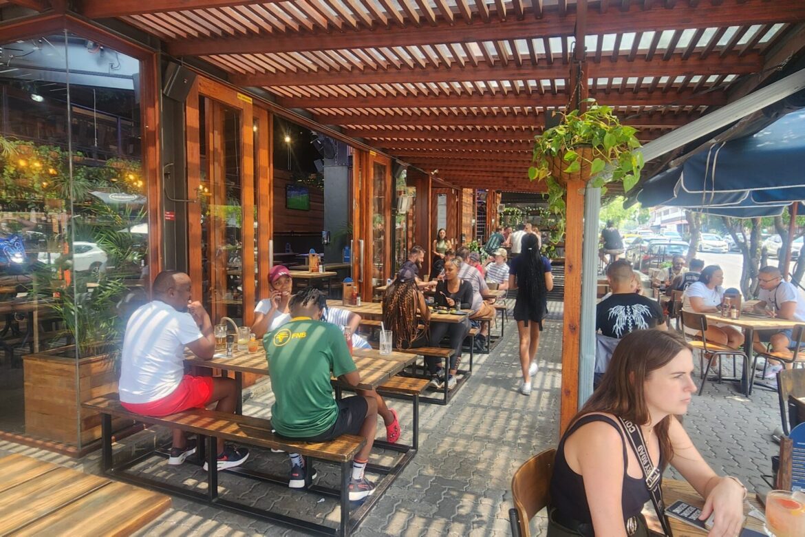 People are sitting at wooden tables on an outdoor patio, shaded by a pergola adorned with lush plants. A few patrons, possibly on a singles food tour, enjoy drinks and conversations. The lively patio is adjacent to a street lined with parked cars.