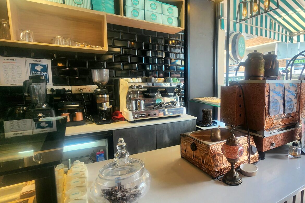 A cozy coffee shop interior invites visitors on a singles food tour, featuring a modern counter and traditional Turkish coffee equipment. The black and white tile backdrop hosts shelves of coffee supplies, while sunlight streams through a window adorned with a green and white striped awning.
