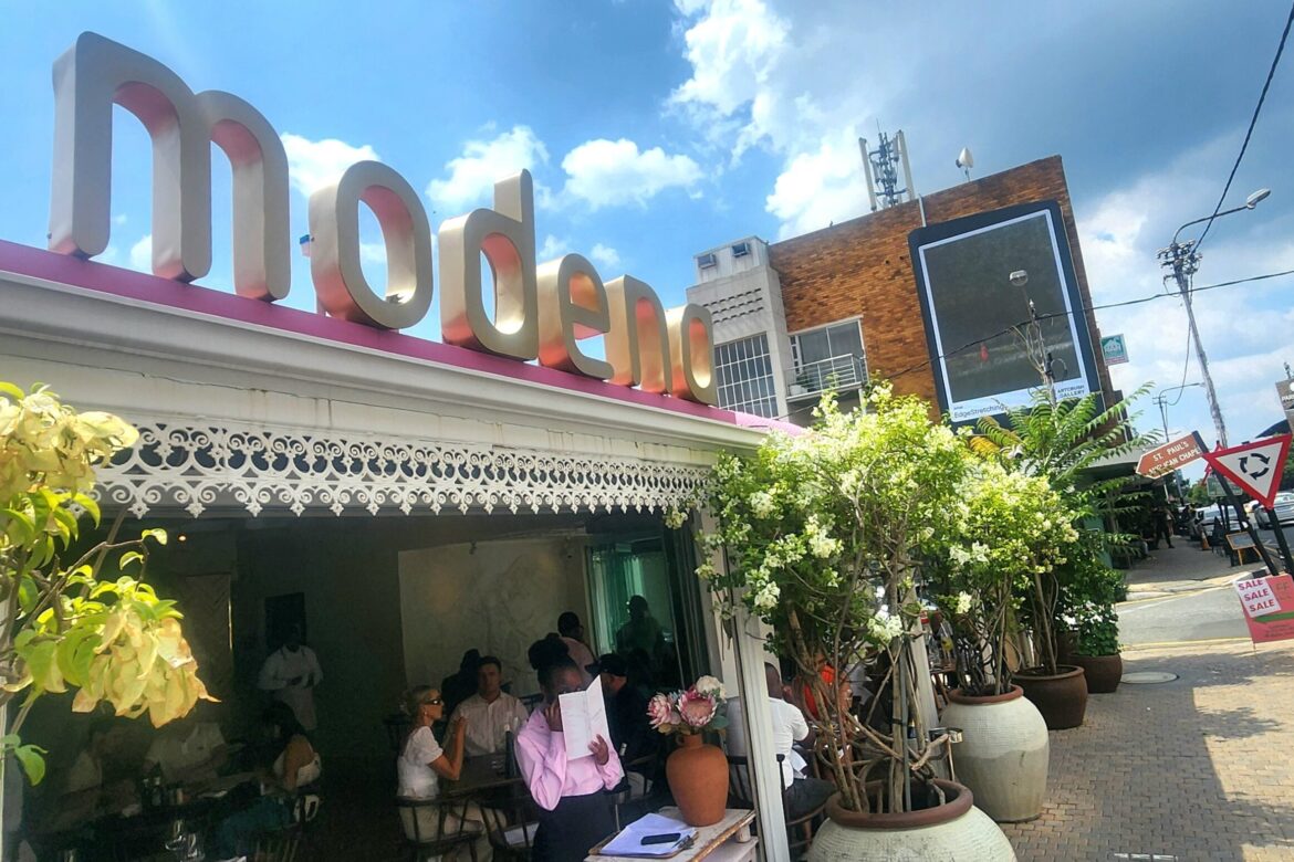 A modern café with a pink and white facade, the word modena gracing its roof, invites visitors on a delightful singles food tour. Inside, patrons enjoy their meals amid potted plants by the entrance. The sunny street reveals other buildings beneath a cloudy sky.