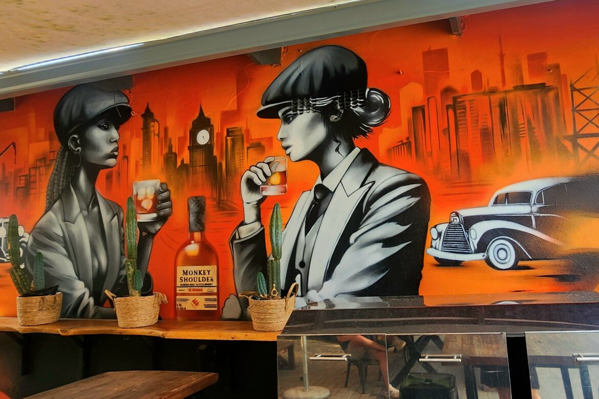 A vibrant mural depicts two stylized figures in vintage attire sipping drinks against an orange cityscape, reminiscent of a classic Singles Food Tour. A classic car is visible in the background, while three small cacti and a bottle of Monkey Shoulder whiskey adorn a shelf beneath.