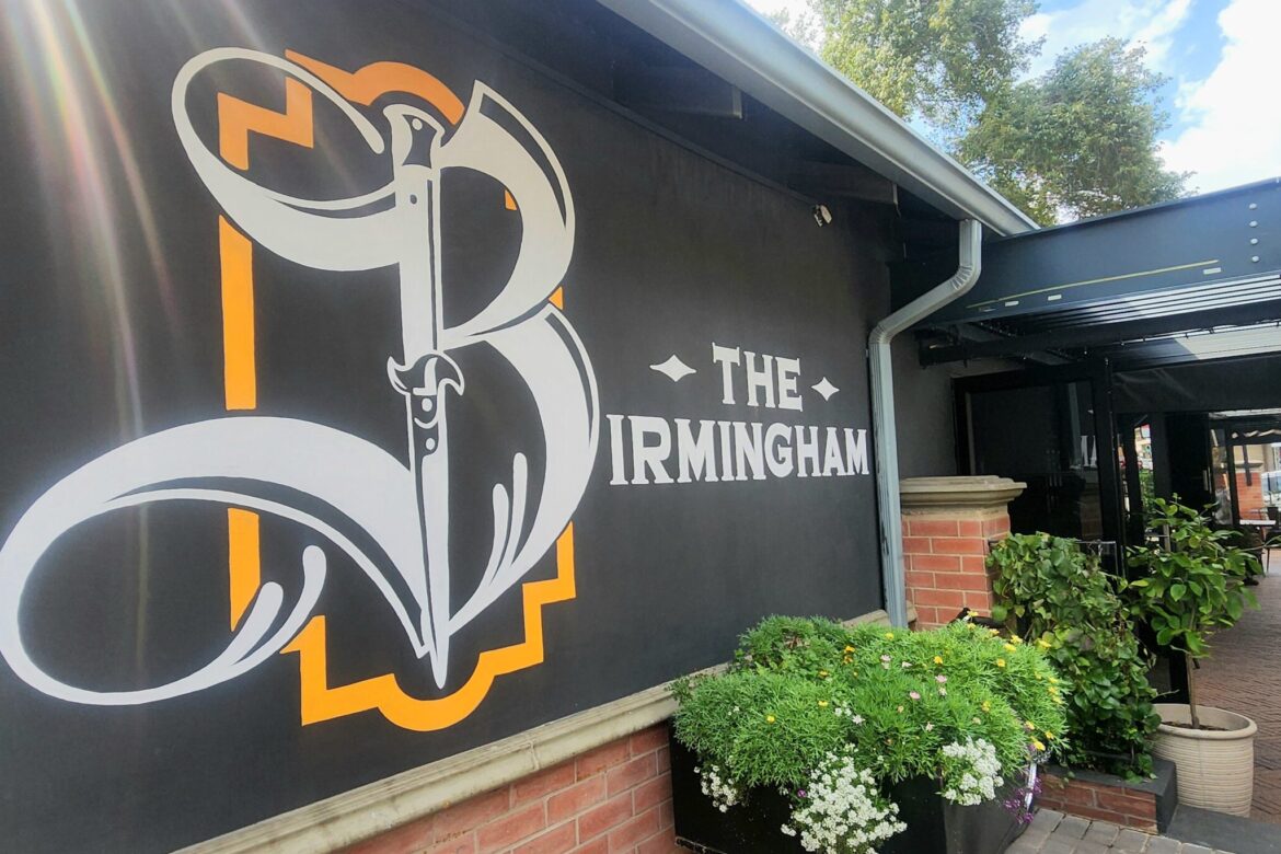 A large sign for The Birmingham adorns a brick building with outdoor seating, perfect for those on a singles food tour. The area is lush with plants and greenery, while a partially covered walkway leads into the distance under a clear blue sky dotted with clouds.